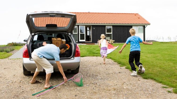 Bildet viser en mann som pakker ut av en bil foran et feriehus. To barn løper inn i huset.