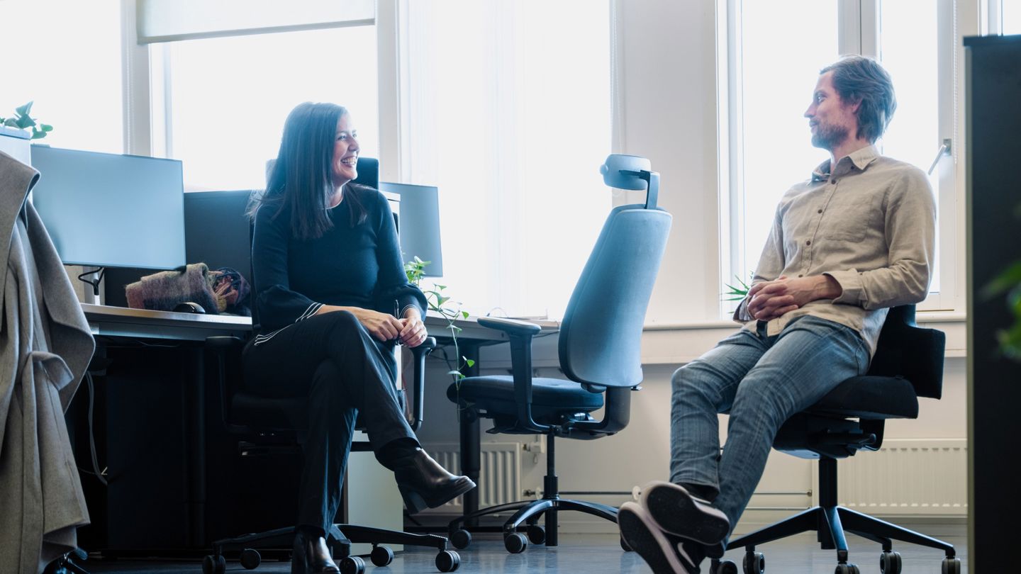 Bildet viser to personer som sitter og snakker med hverandre i et åpent kontorlandskap