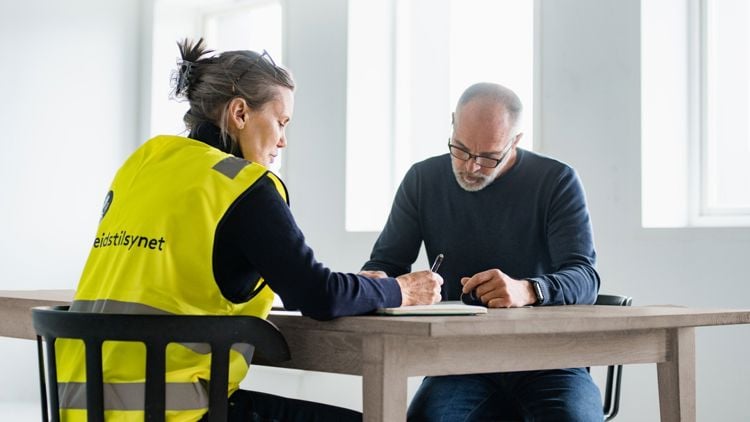 Bildet viser to personer som sitter ved et bord. Den ene har på seg gul Arbeidstilsynet-vest. 