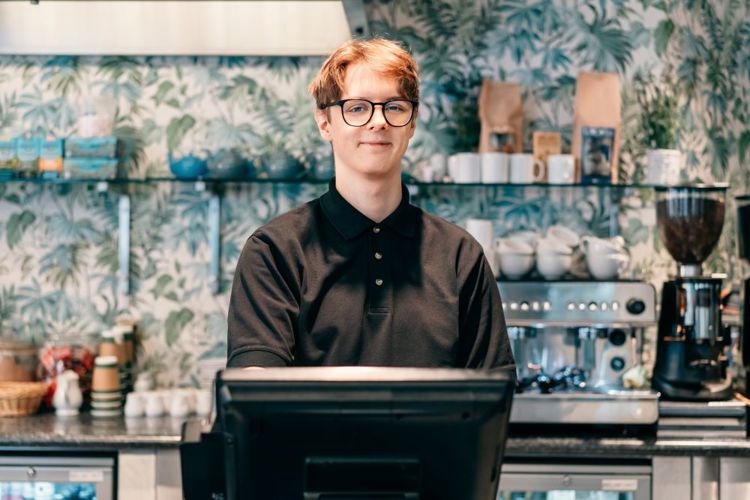 Bildet viser en ung mann som står bak ei kasse på et serveringssted. Mannen kikker rett i kameraet og smiler.