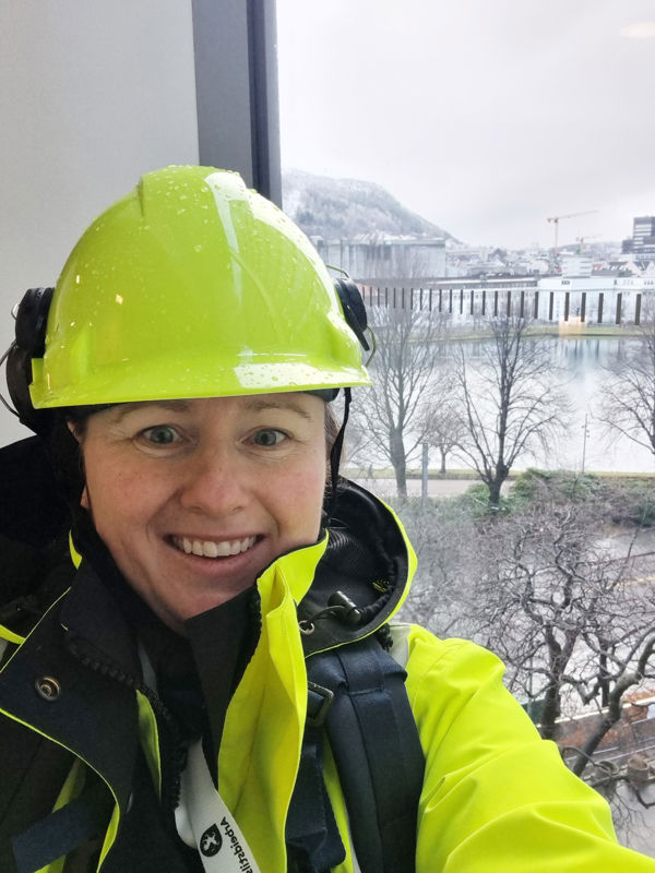 Kvine med gul arbeidshjelm og gul arbeidsjakke foran et vindu med utsikt 