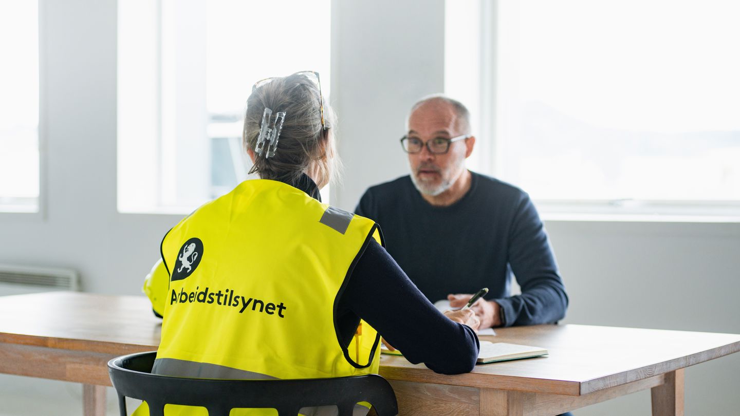 En kvinnelig inspektør i gul vest med Arbeidstilsynet logo sitter ved et bord og snakker med en mann